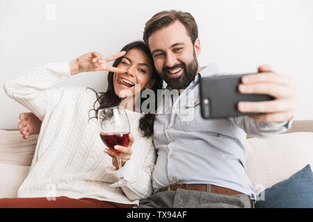Image of positive couple man and woman taking selfie photo together on sofa while drinking wine in bright room at home Stock Photo