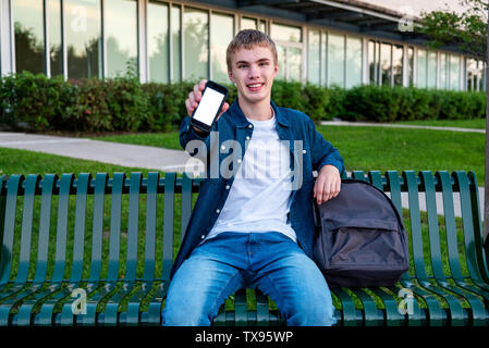 Happy male teenager showing off his phone while sitting on a bench. Stock Photo