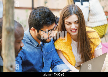 Three multicultural happy smart students looking some files at the laptop while sitting at the outdoor cafe Stock Photo