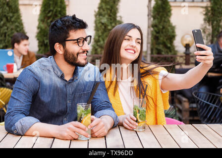 Lovely multicultural couple making selfie at the smartphone while sitting with mohito at the open air cafe Stock Photo
