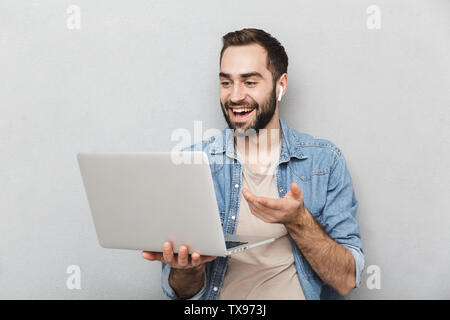 Photo of brunette smiling man having beard holding laptop and using earpod isolated over gray wall Stock Photo