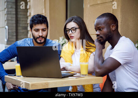 Three multicultural smart students discussing something at the laptop while studying at the cafe Stock Photo