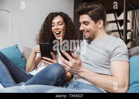 Picture of happy young couple man and woman 30s sitting on sofa at home and using smartphones together Stock Photo