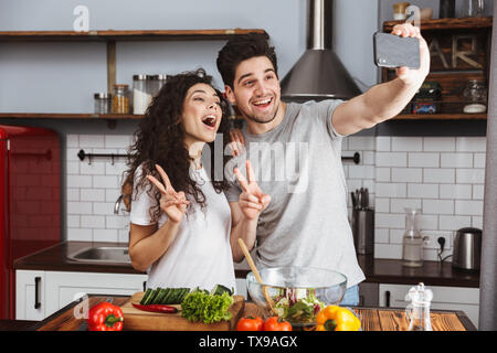 Picture of candid couple man and woman 30s taking selfie photo while cooking salat with vegetables together in modern kitchen at home Stock Photo