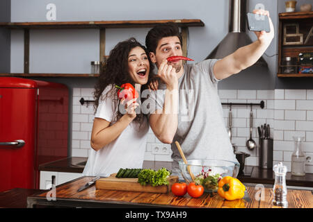 Picture of caucasian couple man and woman 30s taking selfie photo while cooking salat with vegetables together in modern kitchen at home Stock Photo