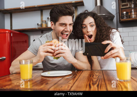 Picture of joyful couple man and woman 30s using mobile phone while eating hamburger during breakfast in kitchen at home Stock Photo