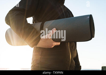Close up of a sportsman wearing jacket walking at the beach, carrying fitness mat Stock Photo