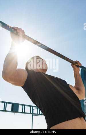 Photo of masculine guy 20s in black tracksuit doing pull ups on horizontal gymnastic bar during morning workout by seaside Stock Photo