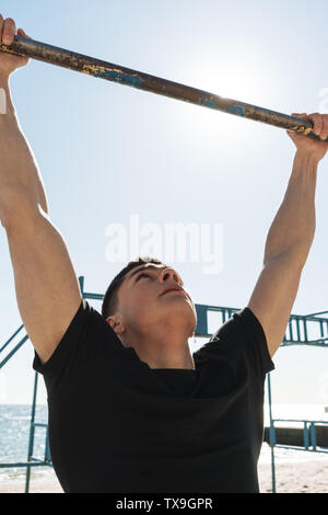 Photo of muscular man 20s in black tracksuit doing pull ups on horizontal gymnastic bar during morning workout by seaside Stock Photo