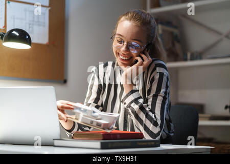 Photo of optimistic caucasian woman architect wearing glasses holding cellphone and house model while sitting at workplace Stock Photo