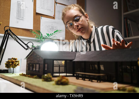 Photo of thoughtful cute woman architect wearing glasses designing draft with house model and sitting at workplace in office Stock Photo