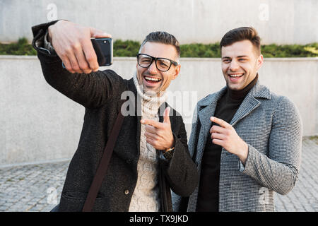 Two handsome men wearing coats spending time outdoors, drinking takeaway coffee, walking, talking, taking a selfie Stock Photo