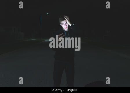 Shocked teenager looking at his phone while standing in the middle of a street at night. His face is being illuminated by the light from the screen of Stock Photo