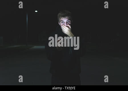 Shocked teenager looking at his phone while standing in the middle of a street at night. His face is being illuminated by the light from the screen of Stock Photo