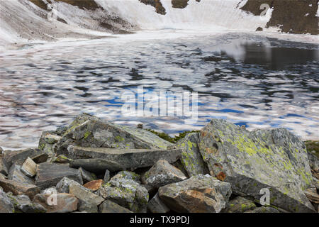 Framed view of The Eye lake on Rila mountain, covered by colorful, cracked ice, by moss covered foreground rocks and background cliff covered by snow Stock Photo