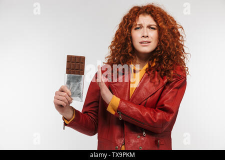 Portrait of healthy redhead woman 20s wearing leather jacket holding chocolate bar and expressing dislike isolated over white background Stock Photo