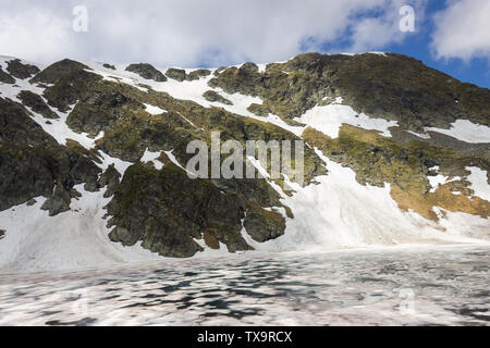 View across The Eye lake, one of the seven Rila lakes, with foreground rocks and colorful, broken ice at a rocky cliff covered by snow Stock Photo