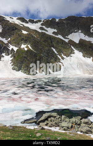 View across The Eye lake, one of the seven Rila lakes, with foreground rocks and colorful, broken ice at a rocky cliff covered by snow Stock Photo