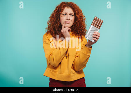 Portrait of displeased redhead woman 20s wearing yellow sweatshirt holding chocolate bar and expressing dislike isolated over blue background Stock Photo