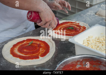 Preparing Pizza Margherita. Pizzaiolo add basil and tomato sauce over a raw pizza dough . Selective focus Stock Photo