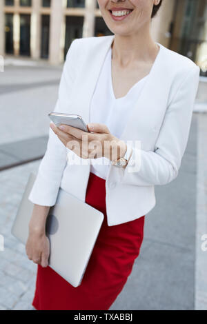 Mid section portrait of smiling businesswoman holding smartphone while posing outdoors in business area, copy space Stock Photo