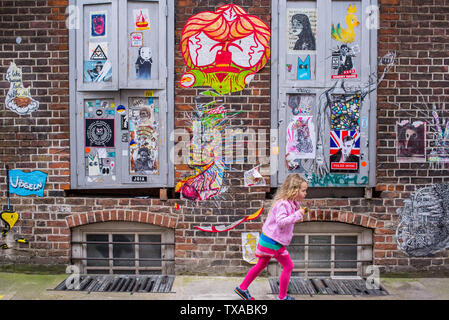 Brick Lane, Shoreditch, London, England, UK: Little young blonde girl running in Brick lane next to a wall covered in graffiti mural street art Stock Photo