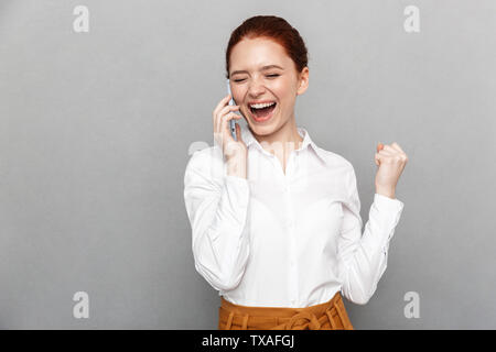 Photo of happy young pretty redhead business woman posing isolated over grey wall background talking by mobile phone make winner gesture. Stock Photo