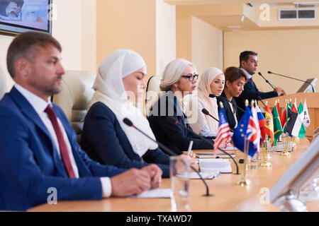 Row of young intercultural delegates in formalwear sitting by table Stock Photo