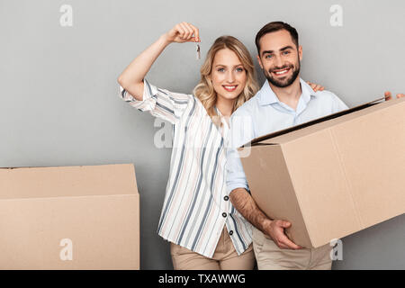 Photo of smiling european couple in casual clothing holding flat keys and carrying cardboard boxes isolated over gray background Stock Photo