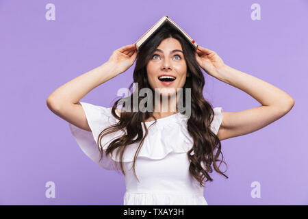 Beautiful cheerful young woman wearing summer outfit standing isolated over violet background, holding diary on her head Stock Photo