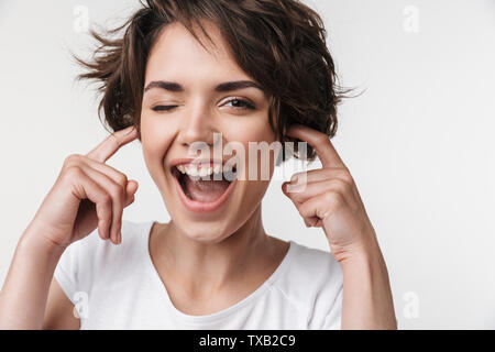 Portrait of optimistic woman with short brown hair in basic t-shirt plugging her ears with fingers isolated over white background Stock Photo