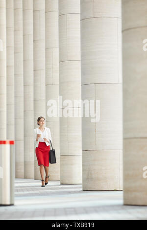 Full length portrait of beautiful woman wearing red skirt walking towards camera along row of pillars, copy space Stock Photo