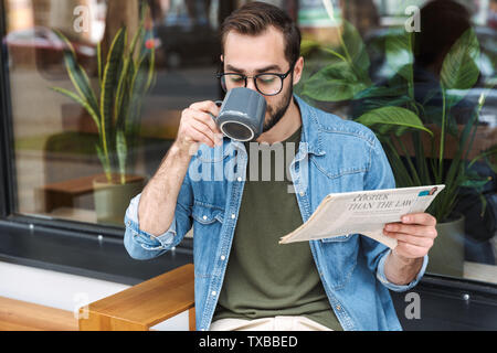 Photo of focused brunette man wearing denim shirt and glasses reading newspaper with cup of coffee in cafe outdoors Stock Photo