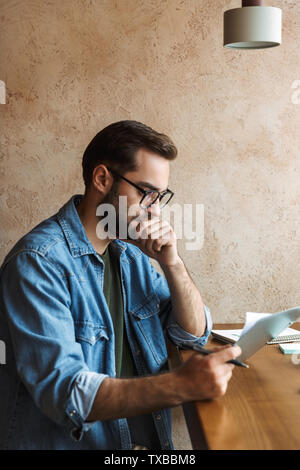 Photo of thoughtful bearded man wearing glasses reading documents while working in cafe indoors Stock Photo
