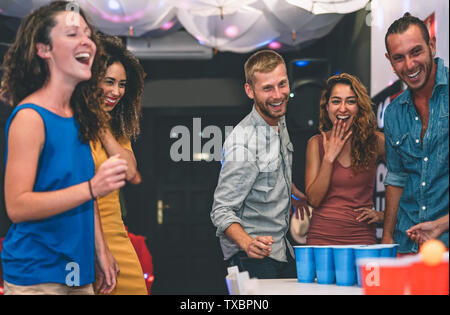 Happy friends playing beer pong in a cocktail bar - Young millennials people having fun doing party alcohol games at night pub Stock Photo