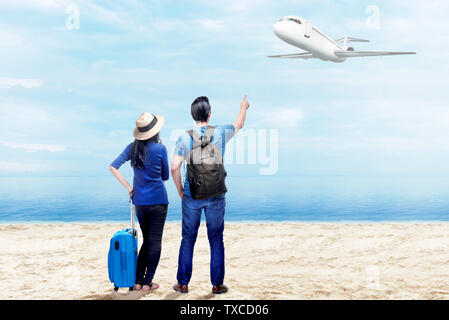 Rear view of asian couple with suitcase bag and backpack standing on the beach and pointing to a flying plane above the sea with blue sky background Stock Photo