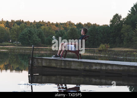 Young man relaxing on an Adirondack chair and looking at a calm river at sunset. Muskoka, Ontario, Canada. Stock Photo