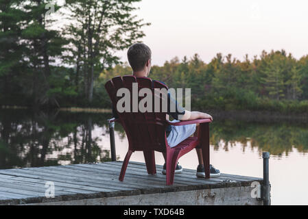 Young man relaxing on an Adirondack chair and looking at a calm river at sunset. Muskoka, Ontario, Canada. Stock Photo