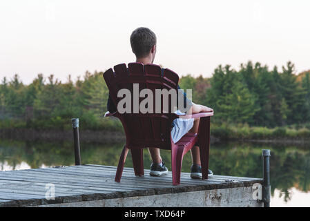 Young man relaxing on an Adirondack chair and looking at a calm river at sunset. Muskoka, Ontario, Canada. Stock Photo