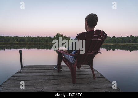 Young man relaxing on an Adirondack chair and looking at a calm river at sunset. Muskoka, Ontario, Canada. Stock Photo