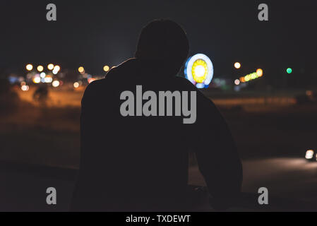 Silhouette of a man leaning on a highway overpass barrier at night. He is looking at the midway of a fair in the distance. Stock Photo
