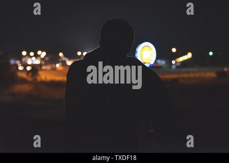 Silhouette of a man leaning on a highway overpass barrier at night. He is looking at the midway of a fair in the distance. Stock Photo