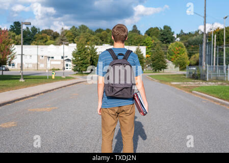 Male teenager with his back turned to the camera and walking towards a school. He is wearing a backpack and carrying some binders. Stock Photo
