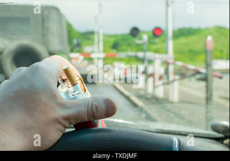 View of the driver hand with a pack of cigarettes on the steering wheel of the car, which stopped before a closed railway crossing at a red light Stock Photo