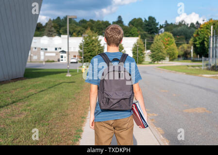 Male teenager with his back turned to the camera and walking towards a school. He is wearing a backpack and carrying some binders. Stock Photo