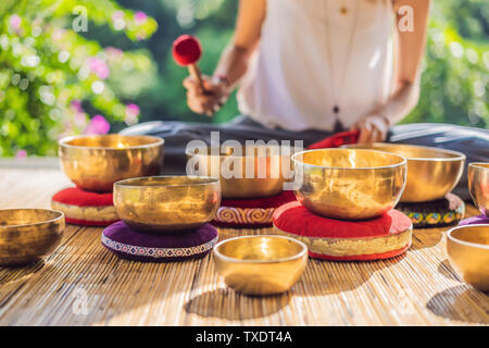 Woman playing on Tibetan singing bowl while sitting on yoga mat against a waterfall. Vintage tonned. Beautiful girl with mala beads meditating Stock Photo