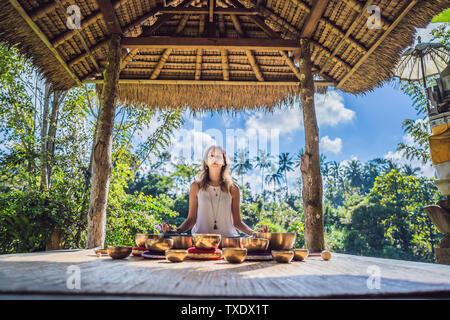 Woman playing on Tibetan singing bowl while sitting on yoga mat against a waterfall. Vintage tonned. Beautiful girl with mala beads meditating Stock Photo