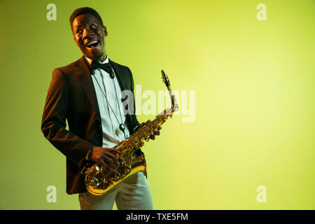 Young african-american jazz musician playing the saxophone on gradient yellow-green studio background. Concept of music, hobby, festival. Joyful attractive guy improvising. Colorful portrait of artist. Stock Photo