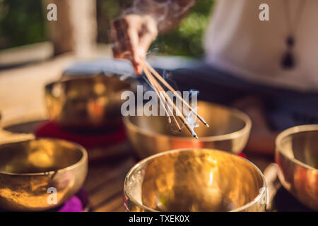 Woman playing on Tibetan singing bowl while sitting on yoga mat against a waterfall. Vintage tonned. Beautiful girl with mala beads meditating Stock Photo