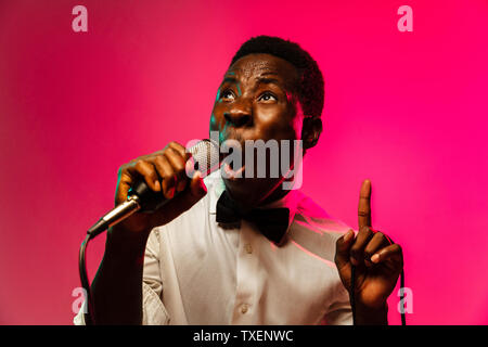 Young african-american jazz musician singing a song on gradient pink-red background in neon light. Concept of music, hobby, festival, open-air. Joyful guy improvising. Colorful retro portrait of singer. Stock Photo